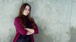 Woman in red plaid shirt leaning against weathered concrete wall with arms crossed.