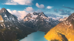 Alpine landscape with turquoise glacial lake nestled between snow-capped mountain peaks under partly cloudy sky.
