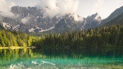 Alpine lake with turquoise water reflecting forested shores and dramatic cloud-shrouded mountain peaks.