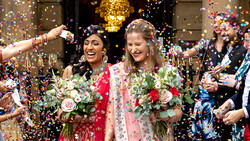 Two people in traditional wedding attire smiling amid falling colorful confetti.