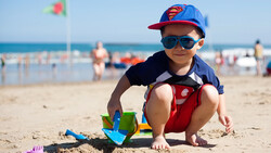 Young child crouching on sand at beach, wearing Superman-themed cap and sunglasses, playing with colorful toy beach equipment.