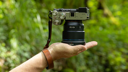 Hand holding a compact mirrorless camera with attached lens against a blurred green outdoor background.