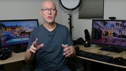 Man in glasses and blue shirt gesturing while speaking in a studio with multiple monitors displaying architectural renderings.