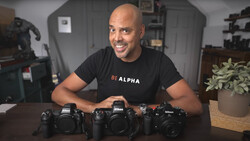 Man in black 'BE ALPHA' shirt displays three professional DSLR cameras on a desk.