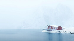 Red houses on a snowy peninsula surrounded by still, misty water in winter conditions.