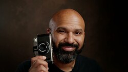 Bearded man holding a vintage film camera up to his face against a dark backdrop.