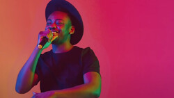Person wearing a black t-shirt and hat singing into a microphone against a gradient background.