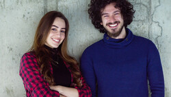 Two smiling people standing against a weathered concrete wall.