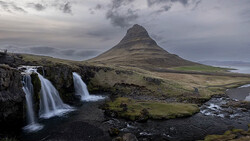 Cone-shaped mountain with cascading waterfalls in foreground under dramatic cloudy sky.