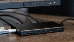 Docking station with multiple ports connected to a laptop, sitting on a wooden desk next to a monitor.