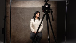 Woman in cream knit sweater seated in studio with camera mounted on tripod and professional lighting setup.