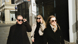 Three people wearing dark coats and sunglasses walking down an urban street.