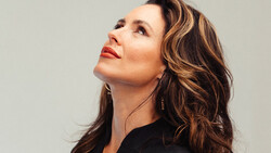 Woman with wavy brunette hair and red lipstick gazing upward against neutral background.