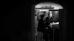 Black and white photograph of two people viewing framed photographs on a gallery wall.