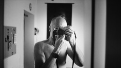 Bald man holding vintage camera up to his face in a mirror self-portrait.