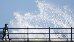 Person in yellow safety vest standing at railing as large wave crashes against barrier.