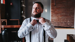 Bearded man in white formal shirt adjusting black bow tie in industrial loft setting.