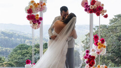 Groom lifting bride in flowing veil with mountain landscape backdrop and hanging floral installations.
