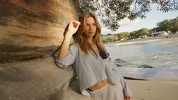 Woman in light blue shirt leaning against weathered wooden post on beach at golden hour.