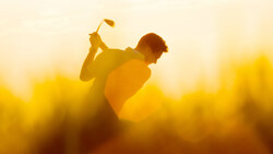 Golfer silhouetted mid-swing against golden sunset sky with blurred background.