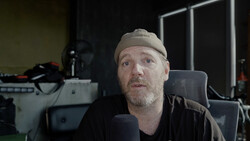 Man wearing a beanie and dark shirt speaking into a microphone in a studio setting.