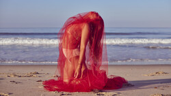 Person wrapped in sheer red fabric on a beach, bent forward with arms extended toward the sand.