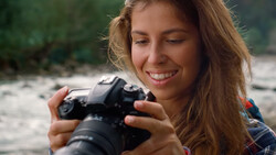 Young woman smiling while holding a DSLR camera with telephoto lens outdoors.