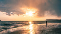 Surfer standing in shallow water during golden hour sunset with dramatic clouds and bright sun breaking through.