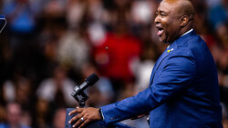 Man in blue blazer speaking at microphone before a crowd.