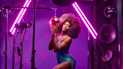 Woman with pink afro holding a disco ball in a neon-lit studio setup with purple lighting.