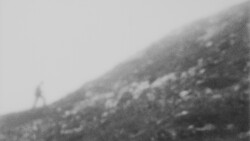 Grainy black and white photograph of a hillside with scattered rocks and sparse vegetation under overcast sky.