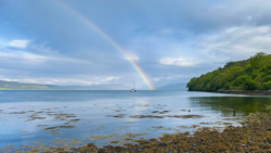 Rainbow stretching from stormy sky to calm bay waters, with forested shoreline and distant sailboat.