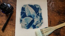 Cyanotype print with blue botanical leaf shapes on white paper, surrounded by art materials on wood surface.