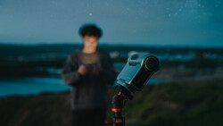 Telescope mounted on tripod pointing toward twilight sky with blurred figure in background.