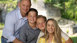 Family of four smiling outdoors in casual clothing with green foliage background.