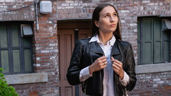 Woman in black leather jacket and white collared shirt standing against brick building backdrop.