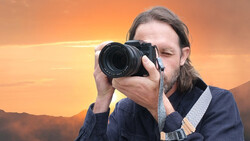 Photographer with long hair and beard holding a telephoto lens camera to his eye against a golden sunset sky.