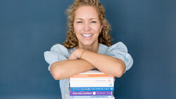 Woman with curly blonde hair smiling at camera while leaning on a stack of colorful books against blue background.