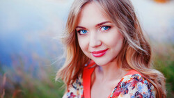 Woman with reddish-brown wavy hair and bright blue eyes, wearing red lipstick and a colorful patterned garment outdoors.