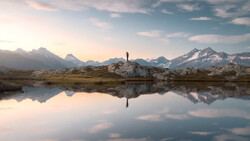Solitary hiker standing on a rocky outcrop, reflected in still water with snow-capped mountains in the background.