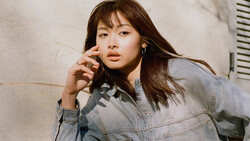Woman in denim jacket with hand near face, shot against textured neutral wall.