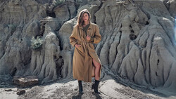 Woman in tan trench coat and black boots standing against dramatic eroded clay badlands formation.