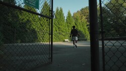 Person running through open gate onto tennis court surrounded by tall green hedges and trees.