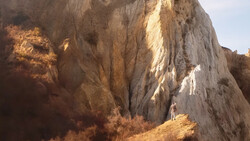 Person standing at the base of a massive eroded cliff face with deep gullies and stratified layers.