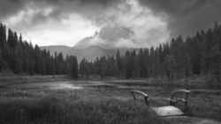 Wooden dock extending into a lake surrounded by evergreen forest with a dramatic mountain peak shrouded in clouds.
