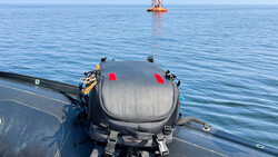 Submersible vessel mounted on a ship deck with offshore platform visible in calm blue water.