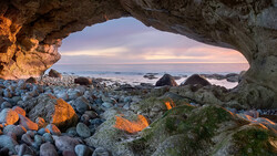 Rocky coastal cave opening framing a twilight seascape with scattered boulders and moss-covered rocks.