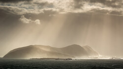 Dramatic island silhouette shrouded in atmospheric mist and volumetric light rays beneath storm clouds.
