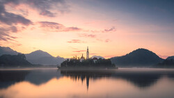 Church with spire on island in calm lake at dawn, surrounded by forested mountains.