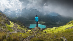 Hiker in blue jacket standing on rocky outcrop overlooking turquoise alpine lake surrounded by misty mountains.